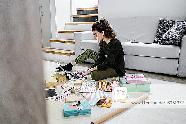 Lächelnde junge Frau sitzt mit eingepackten Geschenken auf dem Boden und benutzt einen Laptop