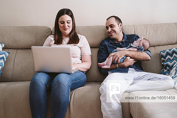 Vater trägt schlafende Tochter  während er die Frau mit dem Laptop auf dem Sofa beobachtet