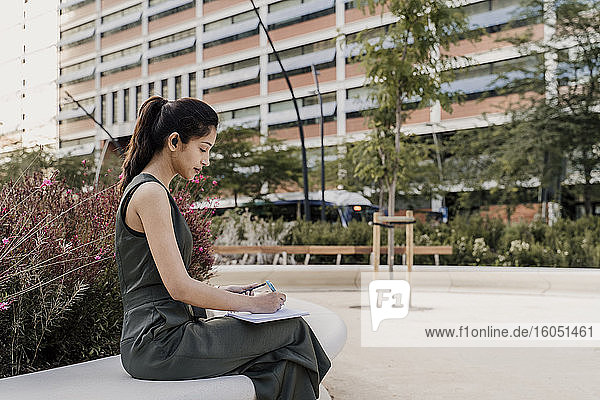 Eine Unternehmerin schreibt in einen Notizblock  während sie auf einem Stuhl vor einem Gebäude sitzt
