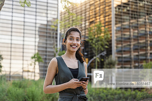 Lächelnde Geschäftsfrau  die ein Mobiltelefon vor modernen Gebäuden in der Stadt benutzt