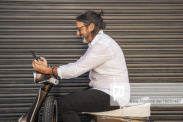 Glücklicher reifer Mann  der sein Smartphone benutzt  während er auf einem Motorroller gegen den Verschluss sitzt