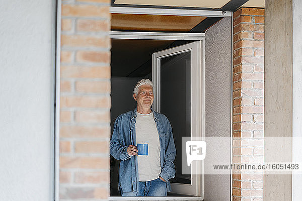 Nachdenklicher älterer Mann  der eine Kaffeetasse hält und durch ein Fenster schaut