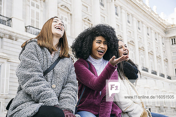 Multi-ethnic female friends laughing while sitting against Madrid Royal Palace  Spain