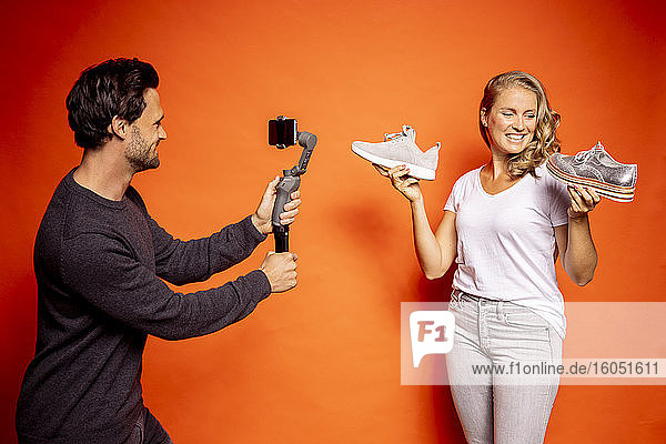 Mann hält Gimbal mit Mobiltelefon  während er eine Frau mit Schuhen vor orangefarbenem Hintergrund fotografiert