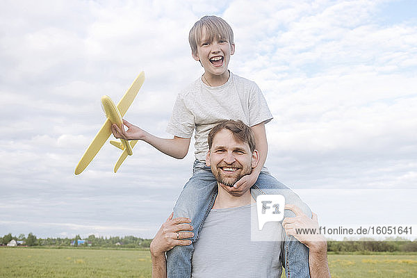 Lächelnder Mann mit Sohn auf den Schultern  der ein Spielzeugflugzeug gegen den Himmel hält