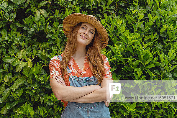 Lächelnde schöne junge rothaarige Frau steht mit verschränkten Armen vor grünen Pflanzen im Hinterhof