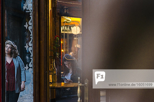 Junge Frau  die ein Smartphone benutzt  während ihre Freundin am Eingang eines Cafés steht  gesehen durch Glas