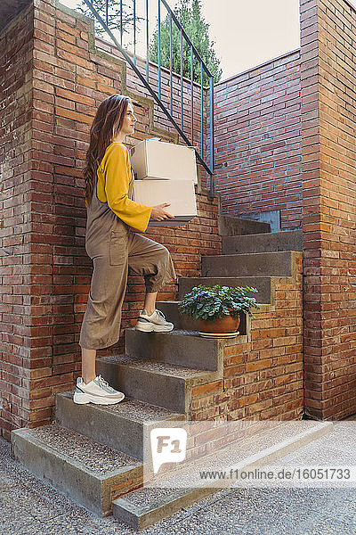 Junge Frau  die weiße Kisten trägt  während sie eine Treppe an einer Backsteinmauer hinaufgeht