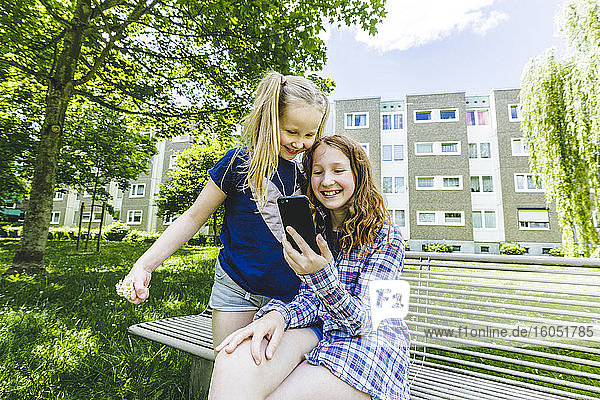 Lächelnde Schwestern teilen sich ein Smartphone auf einer Bank im Park