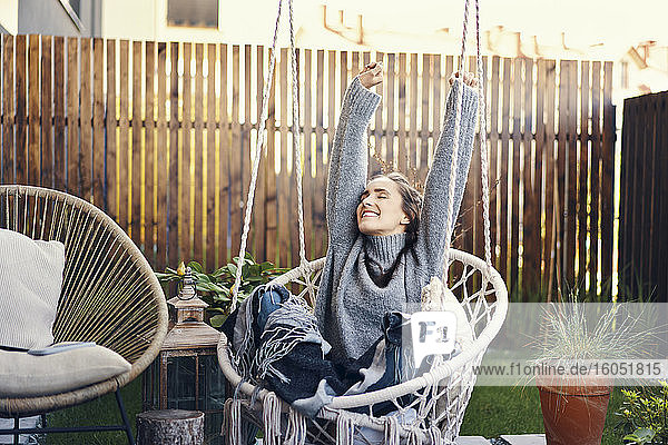 Glückliche junge Frau mit erhobenen Armen  die sich auf einer Schaukel im Garten entspannt