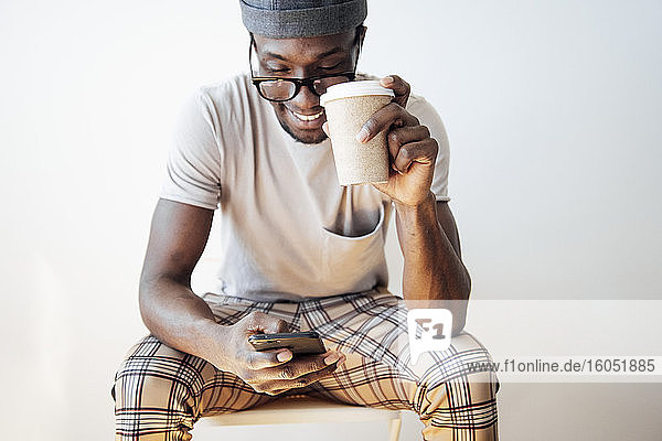 Lächelnder Mann  der eine Textnachricht schreibt  während er eine Kaffeetasse hält und vor einem weißen Hintergrund sitzt