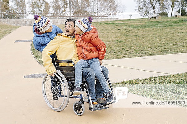 Glücklicher Mann mit aufgeregten Söhnen  die im Rollstuhl auf einem Fußweg im Park fahren
