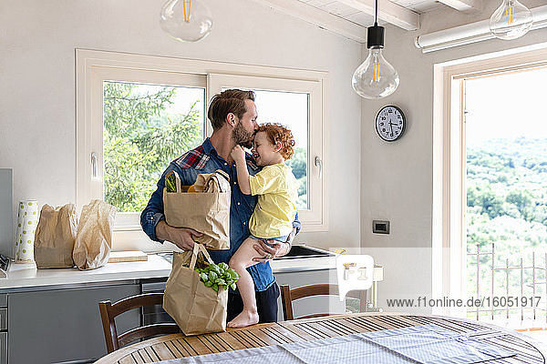 Mann küsst Sohn  während er in der Küche zu Hause die Einkaufstüte hält