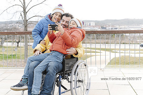 Fröhlicher Junge macht Selfie mit Vater und Bruder im Rollstuhl im Park