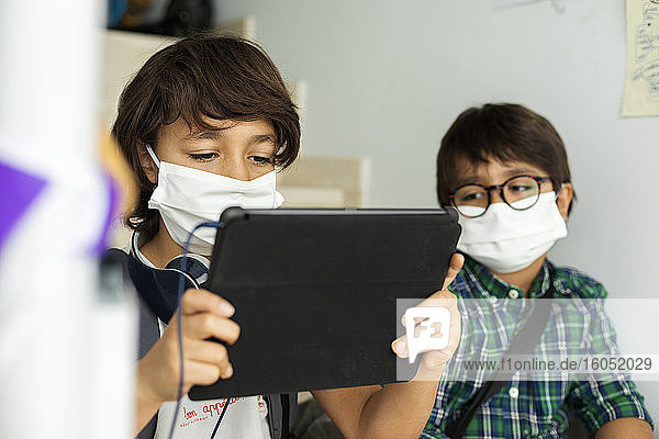 Boy wearing face mask using digital tablet while sitting in distance with friend at school
