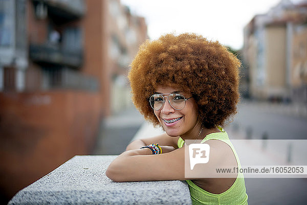 Nahaufnahme einer lächelnden Frau mit Afro-Haar  die an einer Stützmauer in einer Stadt steht