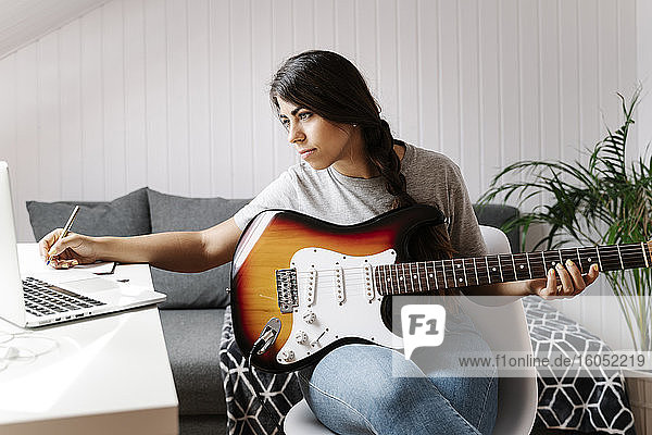 Young woman writing while using laptop and electric guitar at home