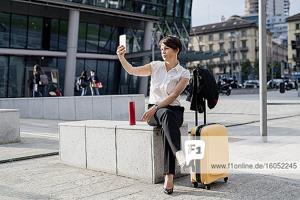 Geschäftsfrau  die ein Selfie mit ihrem Smartphone macht  während sie mit ihrem Koffer auf einem Sitz in der Stadt sitzt