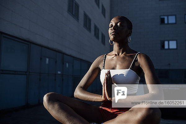 Junge Frau mit geschlossenen Augen meditiert  während sie an einem Gebäude in der Stadt sitzt