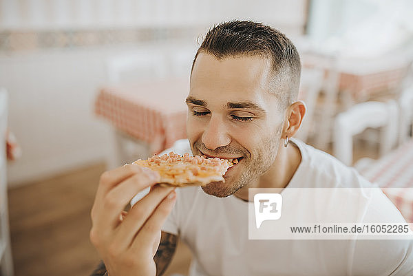 Nahaufnahme eines jungen Mannes  der mit geschlossenen Augen eine Pizza isst  während er im Restaurant sitzt