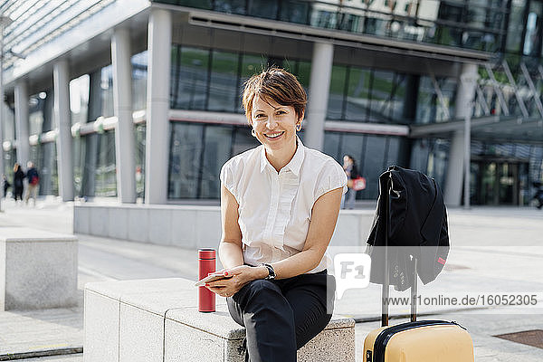 Lächelnde Geschäftsfrau mit kurzen Haaren sitzt auf einem Stuhl vor einem Gebäude in der Stadt