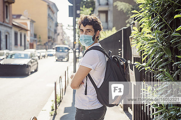 Junger Mann mit Maske und Rucksack steht auf dem Bürgersteig in der Stadt