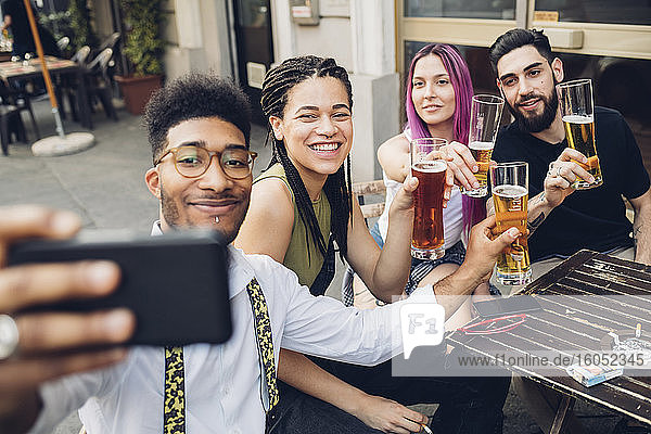 Happy friends holsing beer glasses and taking a selfie outdoors at a bar