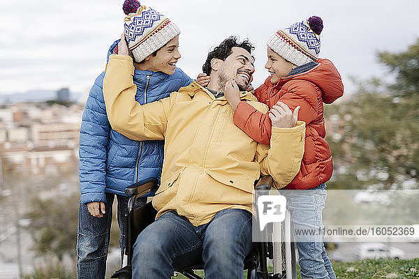 Liebevoller Vater umarmt seine Söhne im Rollstuhl sitzend im Park