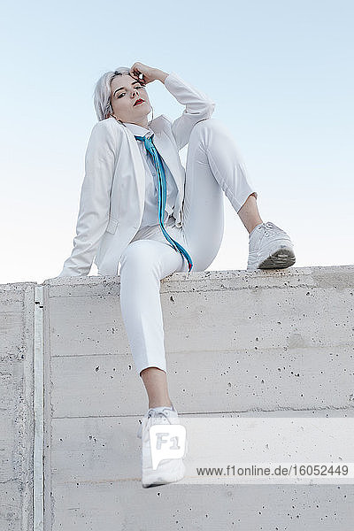 Coole junge Frau im weißen Anzug sitzt auf einer Stützmauer gegen den klaren Himmel