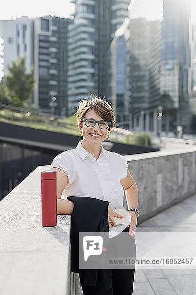 Smiling businesswoman wearing eyeglasses standing by retaining wall in city