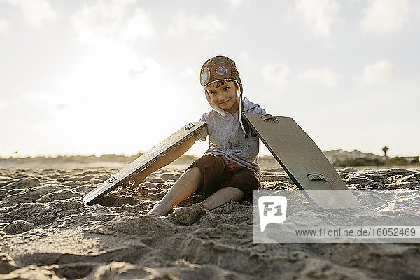 Junge mit Flugzeugflügeln und Mütze auf Sand am Strand sitzend