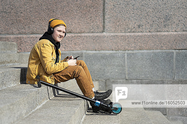Thoughtful boy listening music while sitting with push scooter on steps during sunny day