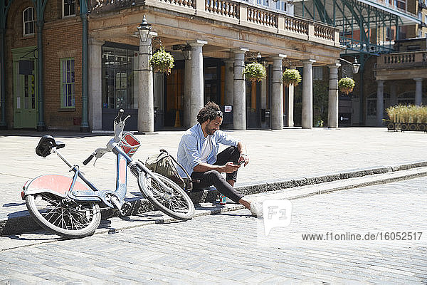 Junger Mann mit Leihfahrrad und Rucksack sitzt am Straßenrand und schaut auf sein Handy  London  Großbritannien