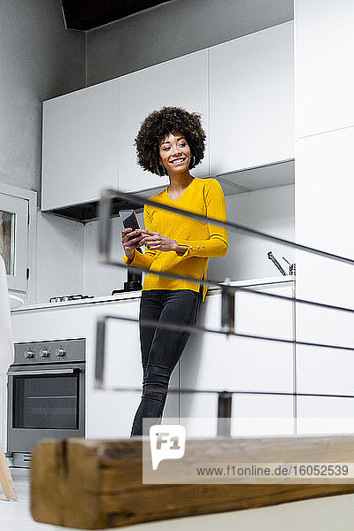 Portrait of smiling young woman standing in the kitchen with smartphone