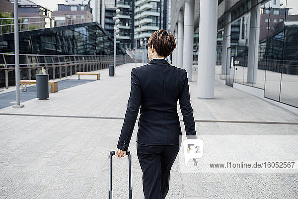 Businesswoman with short hair holding suitcase while walking with footpath in city