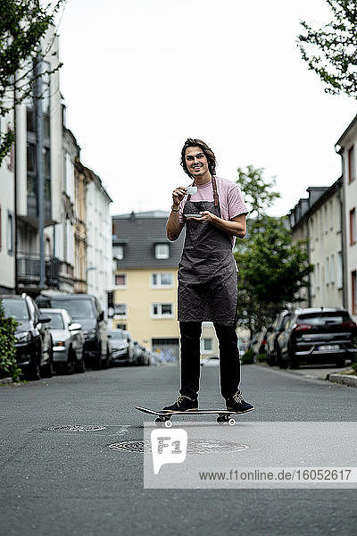 Lächelnder männlicher Besitzer  der eine Kaffeetasse hält  während er auf einer Straße in der Stadt Skateboard fährt