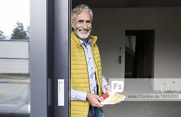 Lächelnder männlicher Architekt hält Farbmuster bei der Renovierung eines Hauses durch ein Fenster gesehen