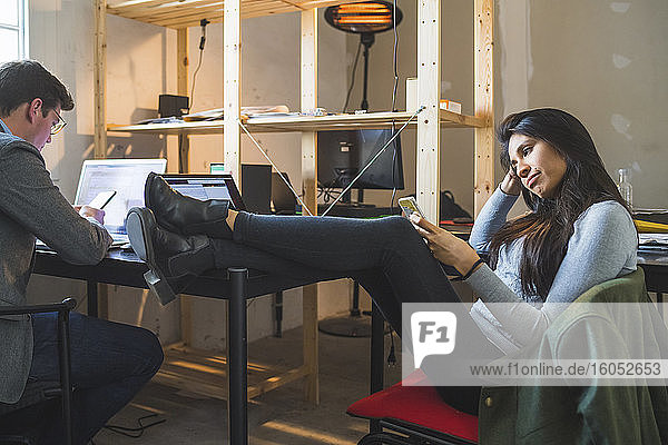 Relaxed woman using smartphone at desk with businessman using laptop
