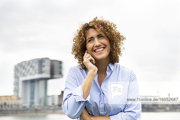 Cheerful female entrepreneur with curly hair talking over smart phone against sky in city