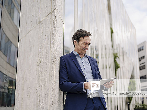 Mature businessman leaning against a building in the city using tablet