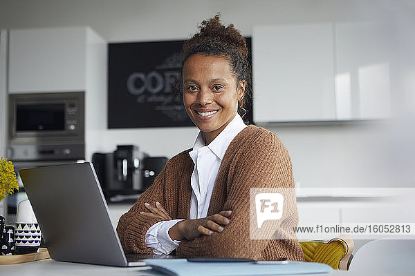 Portrait of smiling businesswoman with laptop sitting at table in kitchen