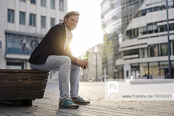 Geschäftsmann sitzt auf einer Bank in der Stadt bei Sonnenuntergang mit einer Flasche Bier