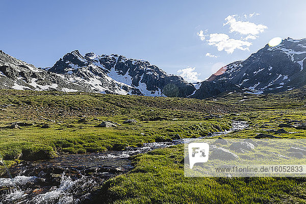 Stream flowing from melting snow near mountains against sky on sunny day