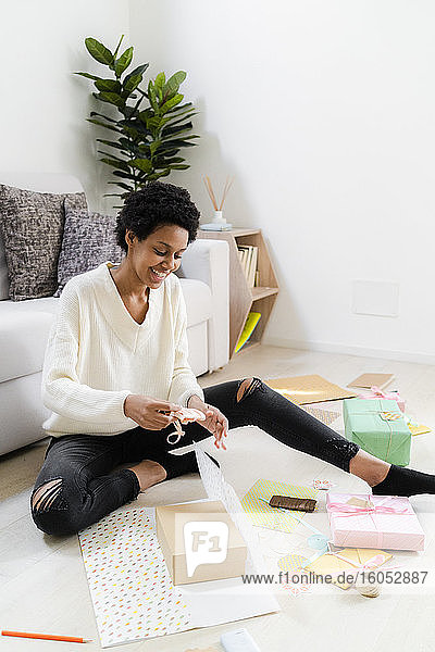 Smiling young woman sitting on the floor at home wrapping presents