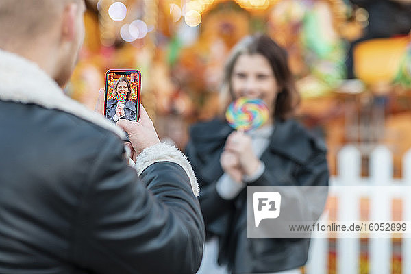 Mann fotografiert Freundin beim Essen eines Lutschers mit dem Smartphone im Vergnügungspark