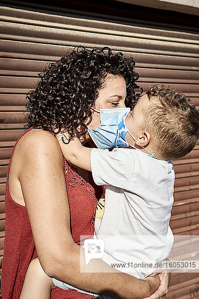 Mutter trägt Sohn mit Masken und küsst ihn  während sie vor einem geschlossenen Fensterladen steht