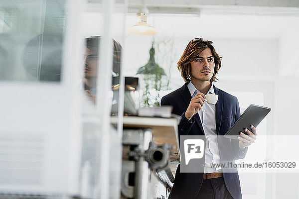 Nachdenklicher männlicher Unternehmer  der ein digitales Tablet und eine Kaffeetasse hält  während er in einem Cafe steht