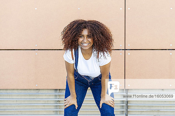 Lächelnde junge Frau mit Afro-Haar  die an einer Wand in der Stadt steht