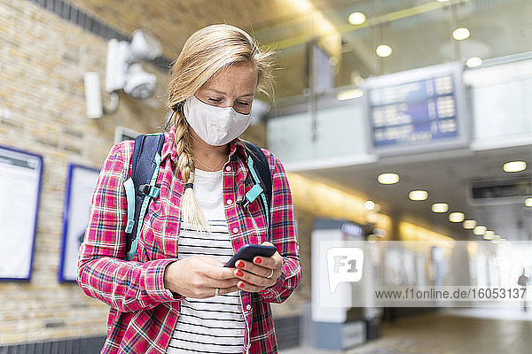 Eine Frau mit Maske benutzt ein Smartphone  während sie an einer U-Bahn-Station steht