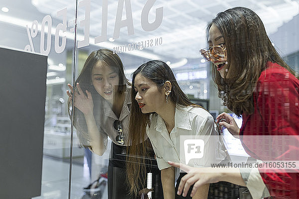 Curious female friends doing window shopping seen through glass in mall